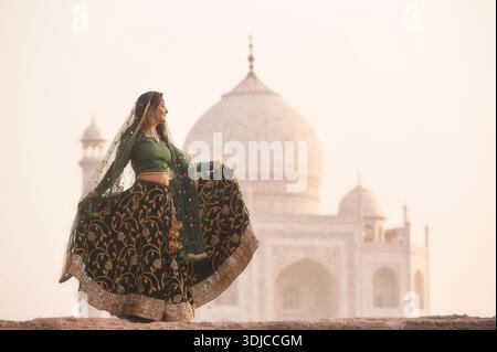 Vue de côté d'une jeune femme dans un sari tenant l'ourlet de sa robe devant le Taj Mahal au coucher du soleil, Agra, Uttar Pradesh, Inde, Banque D'Images