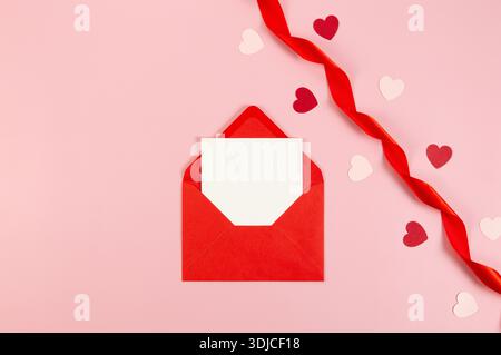 Vue de dessus de l'enveloppe rouge, carte blanche et ruban rouge sur fond rose. Saint Valentin, coeurs rouges et roses, amour. Tissu roulé brillant, soie. Banque D'Images