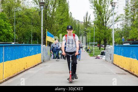 Kiev, Ukraine - 7 mai 2022 : jeune homme avec un sac à dos à cheval sur un trottinette à travers un pont piétonnier avec des clôtures bleues et jaunes à Kiev, Ukraine Banque D'Images