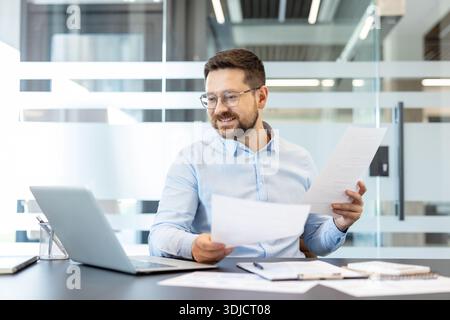 Homme d'affaires barbu professionnel portant des lunettes analysant des documents et examinant des données tout en travaillant sur un ordinateur portable dans un bureau d'entreprise moderne, démontrant la concentration et la productivité de l'entreprise Banque D'Images