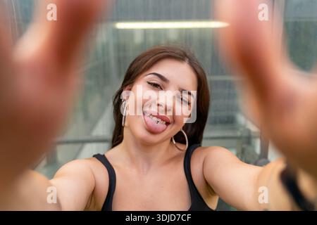 Jeune femme faisant une expression ludique, sortant la langue avec du piercing tout en prenant un selfie en gros plan Banque D'Images