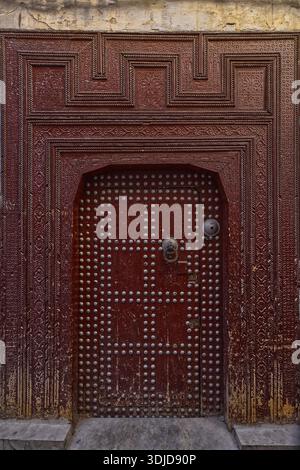 099 porte en bois peinte marron avec décoration cloutée entourée d'un cadre richement sculpté, d'une maison traditionnelle dans la médina el-Bali. Fès-Maroc. Banque D'Images