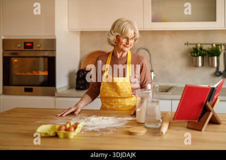 Femme âgée avec de la farine et des œufs sur l'îlot de cuisine lisant le livre de recettes tout en faisant des biscuits à la maison Banque D'Images