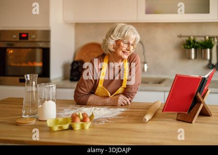 Femme âgée avec de la farine et des œufs sur l'île lisant livre de recettes tout en bloguant sur la cuisson à la maison Banque D'Images