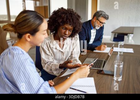Professionnels féminins concentrés lisant des documents tout en étant assis à côté d'un collègue masculin dans la salle de conseil moderne pendant la réunion d'affaires Banque D'Images