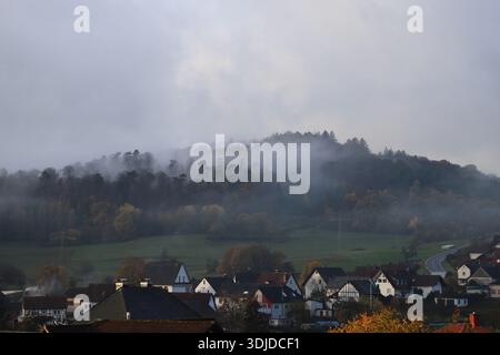 Matin d'automne brumeux dans un petit village européen avec des maisons confortables au premier plan et des collines boisées couvertes de nuages bas et de brouillard. Banque D'Images