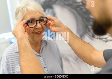 Mains coupées du médecin portant des lunettes à la femme âgée pendant l'examen de la vue à la clinique montrant les soins et le soutien Banque D'Images