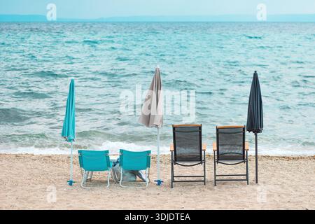 Une scène de plage sereine avec deux paires de chaises longues et des parasols fermés face à la mer turquoise. Le rivage sablonneux est légèrement galbé, et doux wav Banque D'Images