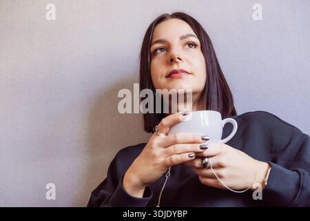Jeune femme aux cheveux foncés tient une tasse de café blanche, assise sur un fond gris, portant un sweat-shirt noir et présentant des ongles manucurés Banque D'Images