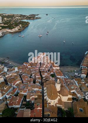 Vue aérienne des toits en terre cuite de la vieille ville descendant en cascade jusqu'à la mer Azur, bateaux parsemant l'eau, Cadaqués, Espagne. Banque D'Images