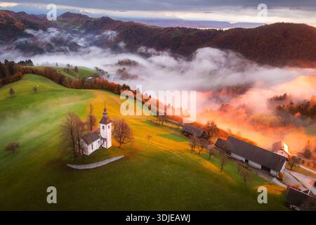 Vue aérienne d'une petite église blanche assise au sommet d'une colline verte alors que la brume monte à travers la vallée, Jamnik, Slovénie. Banque D'Images