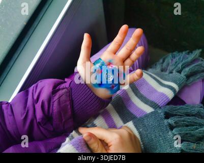 Gros plan de la paume ouverte d'un enfant affichant une montre-jouet LED bleue qui tourne. Le sujet porte une veste d'hiver violette et une écharpe rayée pendant son voyage. Banque D'Images