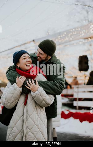 Une coréenne rit alors qu'un homme caucasien l'embrasse par derrière à un festival d'hiver. Ils sont entourés de lumières et de décorations festives. Th Banque D'Images