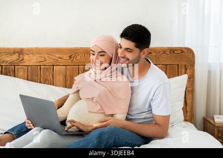 Joyeux couple arabe enceinte utilisant un ordinateur portable, regardant le film ensemble tout en se reposant sur le lit Banque D'Images