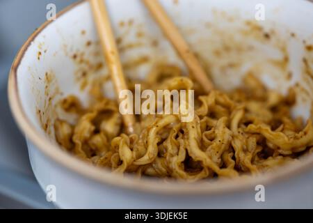 Plan rapproché et en grand angle d'un bol en céramique blanche contenant des nouilles ramen bouclées cuites enrobées d'une sauce brune savoureuse. Deux baguettes en bois reposent i. Banque D'Images