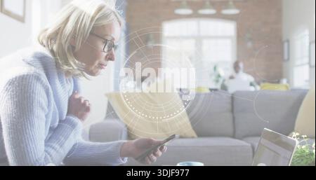 Tenant le téléphone, femme mature avec des lunettes, pull penché dans le salon avec ordinateur portable, canapé gris, HUD Banque D'Images