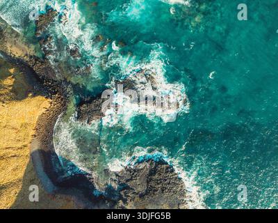 Les vagues turquoises vibrantes de l'océan s'écrasent contre une formation rocheuse sombre et accidentée et une côte texturée brun doré d'une perspective aérienne. Banque D'Images