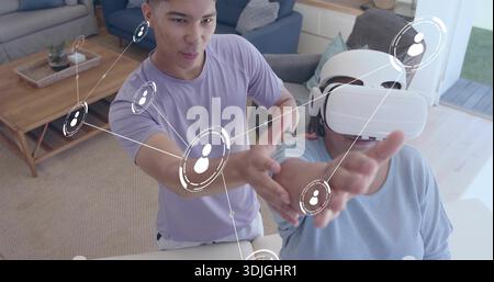Homme adulte guidant une femme senior portant un casque VR blanc dans le salon à la maison, avec des graphiques AR Banque D'Images