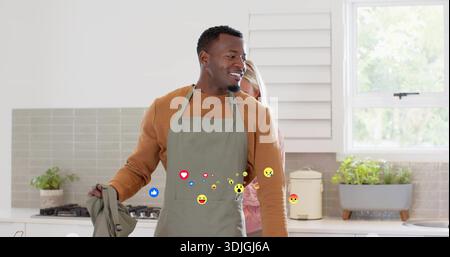 Homme noir debout portant un chandail de rouille gris tablier tenant une serviette souriante dans la cuisine, avec emojis Banque D'Images