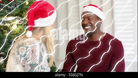 Couple souriant portant des chapeaux de Père Noël et des chandails, assis face à la maison avec arbre de Noël, superposition Banque D'Images