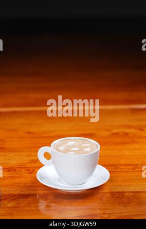 Café chaud fraîchement préparé avec de la mousse légère, servi dans une tasse en céramique blanche et soucoupe sur une table en bois poli. Banque D'Images