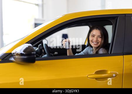 Bonne brune femme achetant une nouvelle voiture mignonne, levant la main avec la clé automatique et souriant, salle d'exposition intérieur, espace de copie. Une cliente plutôt curieuse, pu Banque D'Images