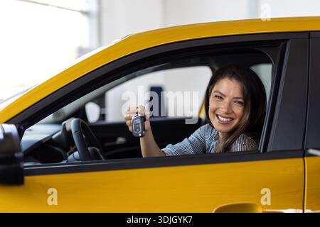 Bonne brune femme achetant une nouvelle voiture mignonne, levant la main avec la clé automatique et souriant, salle d'exposition intérieur, espace de copie. Une cliente plutôt curieuse, pu Banque D'Images