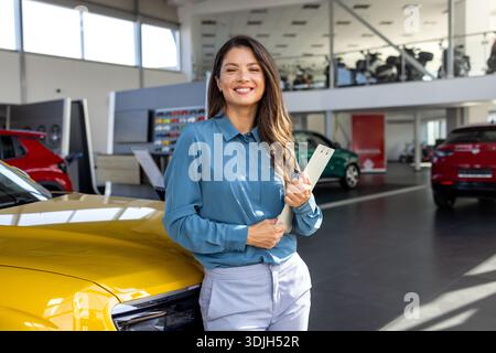 Femme caucasienne en tenue professionnelle tenant des documents, debout à côté d'un modèle de voiture nouvellement lancé dans une concession élégante, mettant en vedette le top automotiv Banque D'Images