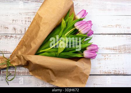 Un bouquet de tulipes hollandaises roses enveloppées dans un papier recyclé brun avec un ruban vert, sur une vieille table peinte en blanc ébréché Banque D'Images