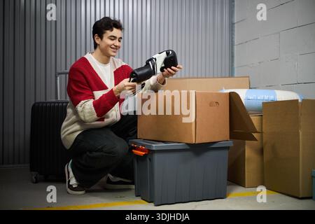 Jeune homme caucasien adulte agenouillé sur le sol déballant la boîte en carton, tenant des gants noirs et de boxe, entouré de conteneurs en plastique et valise dans l'unité de stockage Banque D'Images