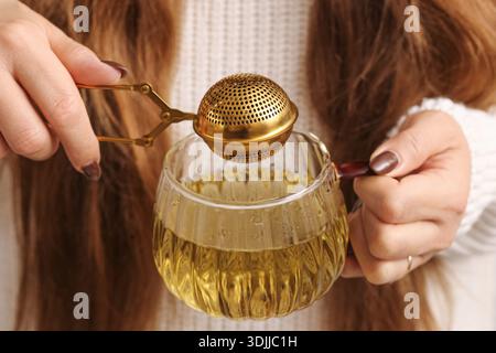 Infusion de thé avec infuseur métallique gros plan. Préparation d'une boisson chaude à base de plantes. Mains féminines faisant du thé. Concept de rituel de boisson maison confortable. Banque D'Images