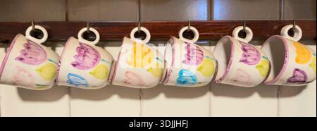 Tasses en céramique peintes à la main avec des motifs de tulipes colorés, suspendues à l'envers sur un support en bois dans une cuisine, représentant une décoration intérieure confortable et buvée Banque D'Images