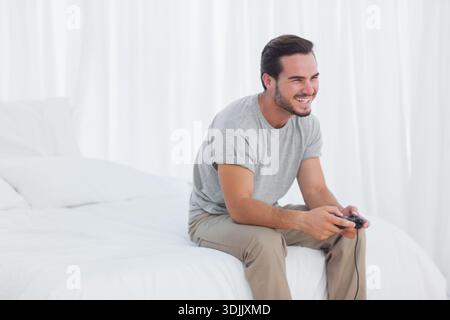Homme assis sur le bord du lit tenant contrôleur filaire souriant et regardant droit dans la chambre, espace de copie. Minimaliste, lumineux, blanc, style de vie, jeux, gamer Banque D'Images