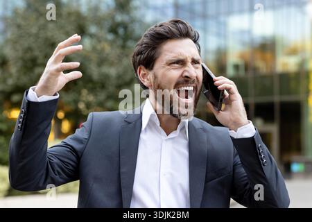 Homme d'affaires en costume éprouvant une frustration et une colère intenses tout en criant dans un smartphone, en faisant des gestes avec une main ouverte, en transmettant le stress et les problèmes de communication à l'extérieur Banque D'Images