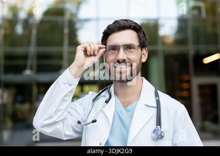 Médecin masculin expérimenté portant un manteau blanc et un stéthoscope, souriant avec confiance et ajustant les lunettes, debout à l'extérieur contre un hôpital moderne ou un bâtiment clinique Banque D'Images