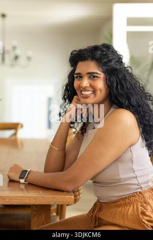 Femme indienne assise à la table à manger en bois penchée sur la main, souriant tout en portant smartwatch Banque D'Images