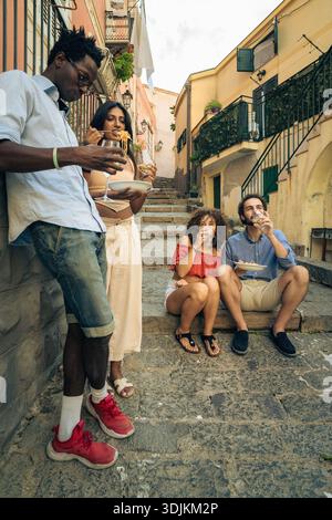 Groupe multiculturel d'amis mangeant des pâtes et buvant du vin ensemble à l'extérieur sur des marches de pierre en Italie. Style de vie authentique de vacances d'été avec trav Banque D'Images