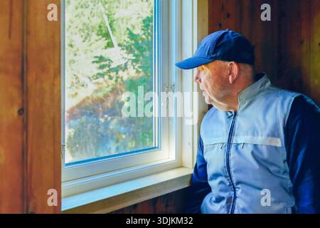 Homme en casquette bleue et veste regarde par la fenêtre, entouré de murs en bois, capturant un moment de réflexion et de connexion avec la nature et tranquilles Banque D'Images