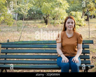 Portrait d'une femme assise sur un banc de parc, dans un environnement verdoyant avec un fond flou et une lumière naturelle. Idéal pour les sujets de style de vie, loisirs Banque D'Images