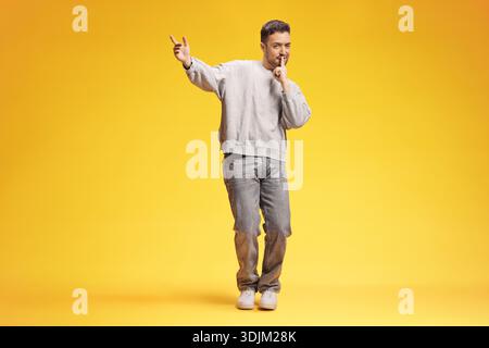 Homme ludique en sweat-shirt gris et Jean faisant un geste tranquille isolé sur fond jaune Banque D'Images