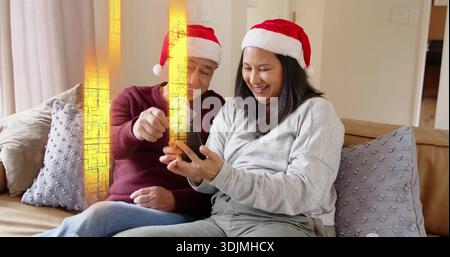 Femme mature souriante et homme senior pointant au smartphone sur le canapé à la maison, portant des chapeaux de Père Noël. Couple, téléphone, festif, confortable, salon, chaleur, éclairage Banque D'Images