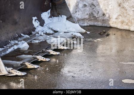 Gros plan des dents de godet de pelle industrielle déblayant la neige gelée et la glace d'une chaussée d'asphalte humide Banque D'Images