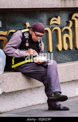 Bangkok, Thaïlande, 19 novembre 2025, un vieil homme de la région est assis tranquillement sur une corniche de pierre, savourant sa collation dans un cadre vibrant de letteri thaïlandais Banque D'Images