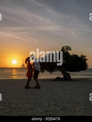 Un couple partage un moment tendre alors que le soleil se couche sur Eagle Beach à Aruba, projetant des teintes dorées sur le rivage serein. La silhouette capture un Banque D'Images