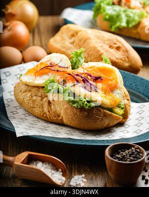 Savoureux sandwich au petit déjeuner composé d'un œuf frit, de laitue et d'autres ingrédients frais sur un petit pain croustillant, servi sur une table en bois avec des condiments Banque D'Images