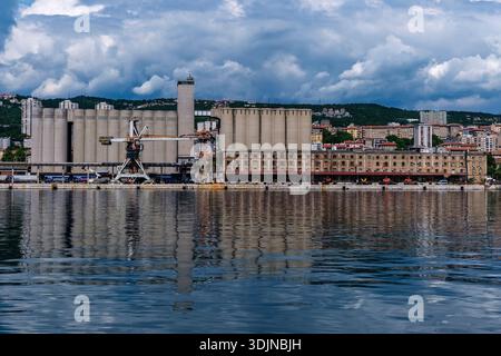 Grands silos à grains et entrepôts portuaires au port de Rijeka. Les bâtiments résidentiels et les collines sont visibles au loin sous un ciel nuageux. Banque D'Images
