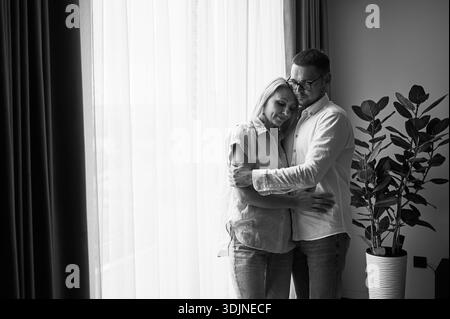 Couple heureux embrassant sur fond clair à la maison. Jolie femme souriant et embrassant son mari les yeux fermés. Couple doux en vêtements décontractés. Image en noir et blanc. Banque D'Images