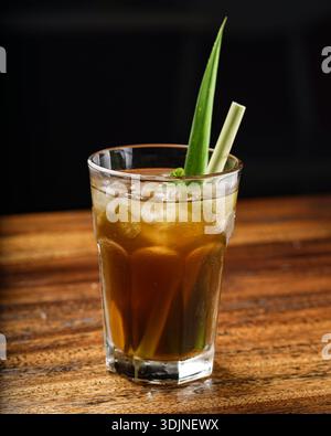 Un grand verre rempli de tisane glacée, garni de menthe fraîche et de tiges de citronnelle. La boisson de couleur ambre est servie sur un onglet en bois rustique Banque D'Images