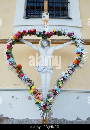 Un crucifix en bois avec une figure peinte de Jésus-Christ, encadré par une grande couronne en forme de coeur de fleurs artificielles colorées sur un mur d'église. Banque D'Images