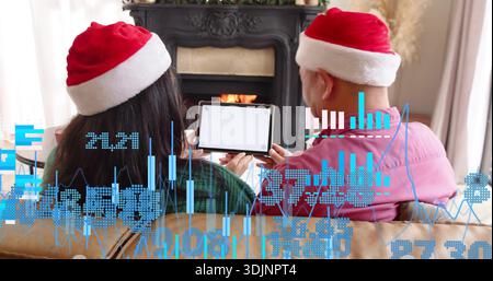 Assis couple dans des chapeaux de Père Noël face à la cheminée, une verte, une rose tenant tablette affichant des graphiques Banque D'Images